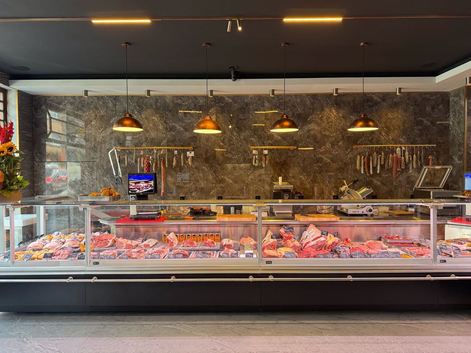 Le comptoir de la boucherie : la longue vitrine réfrigérée pleine de viandes, le mur en marbre sombre et les suspensions en cuivre